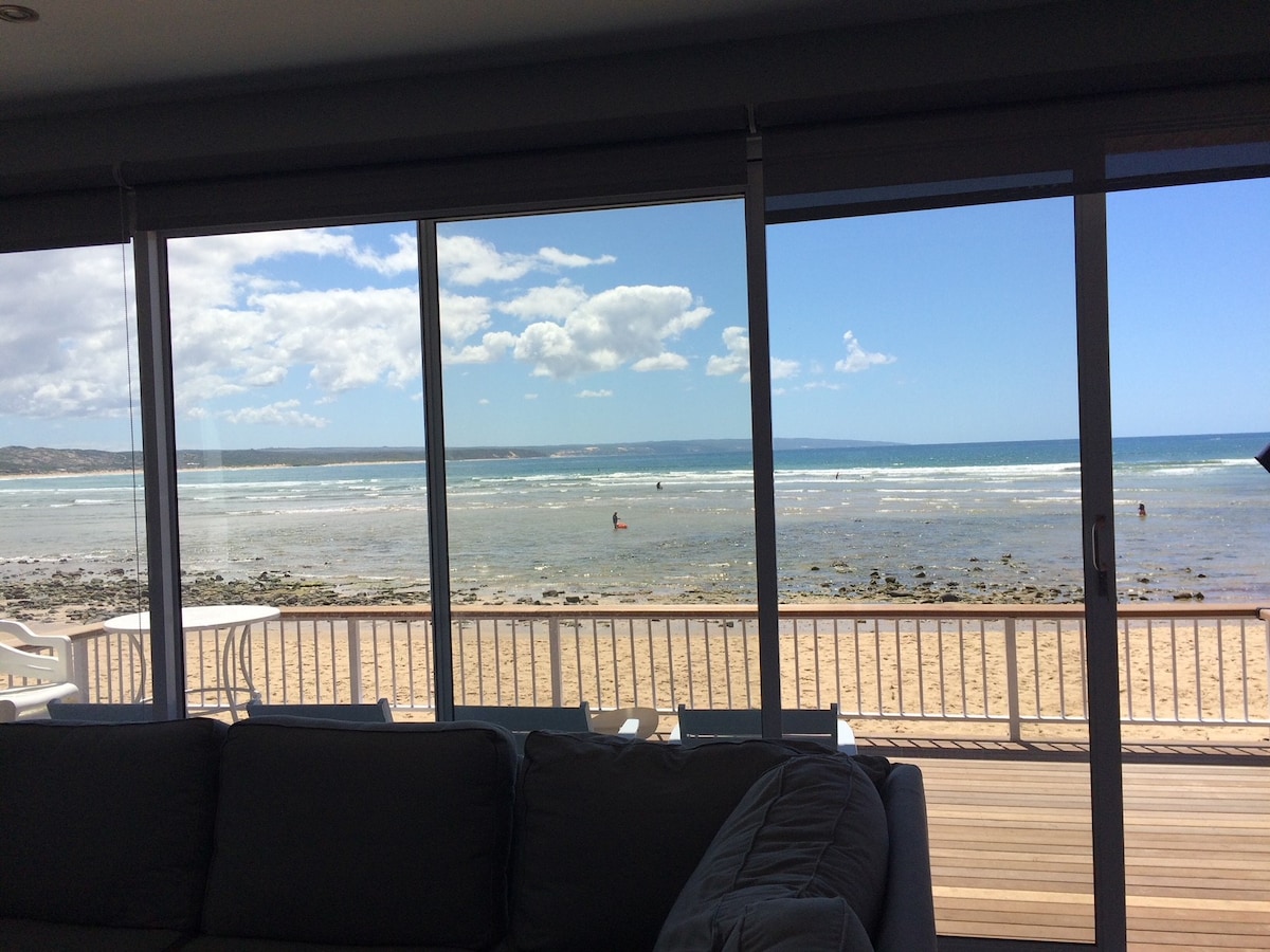 A panoramic view is captured through large glass doors, revealing an ocean scene with gentle waves lapping against the shore. The sandy beach extends in front, while people can be seen enjoying the water. Sunlight brightens the space, reflecting off the glass.