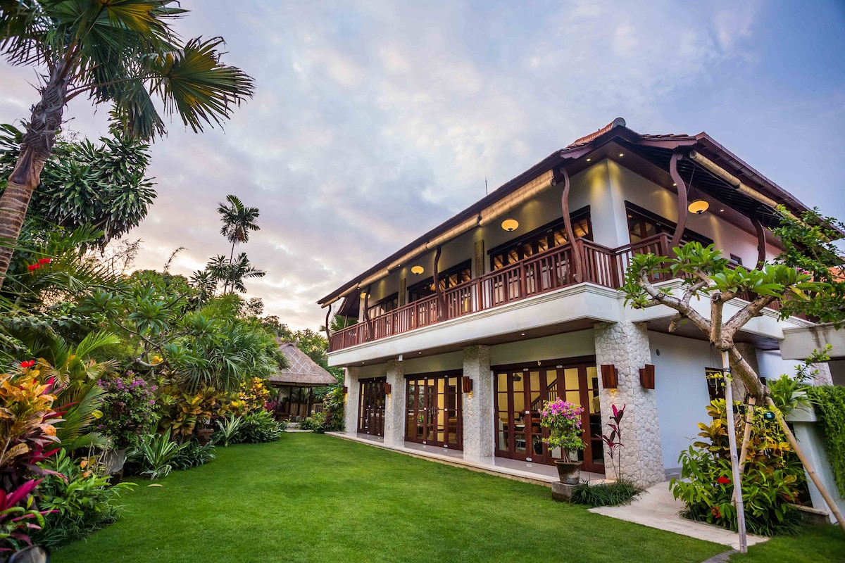 The villa's exterior features a two-story structure adorned with intricate architecture, surrounded by lush tropical gardens. A spacious lawn stretches out, framed by vibrant plants and trees. Large glass doors invite natural light, enhancing the serene atmosphere.