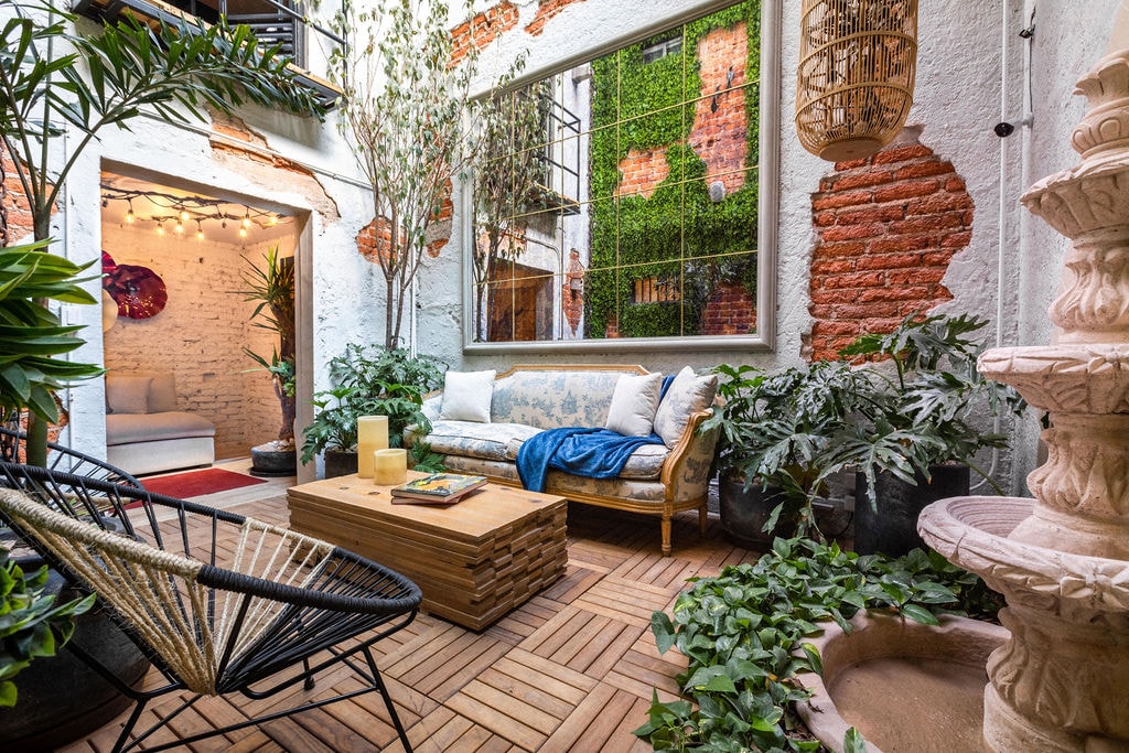 A tranquil courtyard features a natural stone fountain, surrounded by lush greenery and decorative plants. A wooden coffee table sits atop a patterned wooden floor, accompanied by a sofa adorned with a blue throw blanket. A large mirror reflects the surrounding plants, enhancing the serene atmosphere.