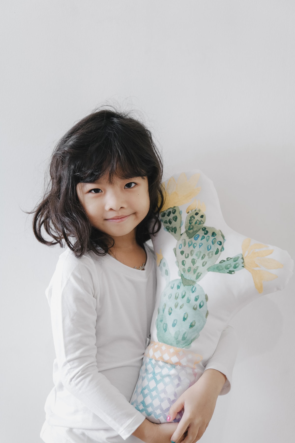 A child stands against a plain white wall, holding a cactus-shaped pillow adorned with floral patterns. The child wears a simple white long-sleeve shirt. The overall setting appears calm and minimalistic, drawing attention to the soft textures of the pillow.