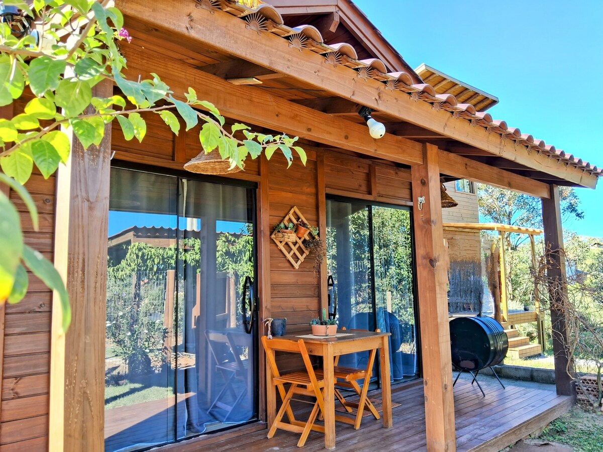 A wooden chalet is featured, showcasing a covered porch with a small dining table and two chairs. Large glass doors provide access to the interior, while lush greenery surrounds the property, enhancing the serene atmosphere. Natural light is evident, creating a welcoming environment.