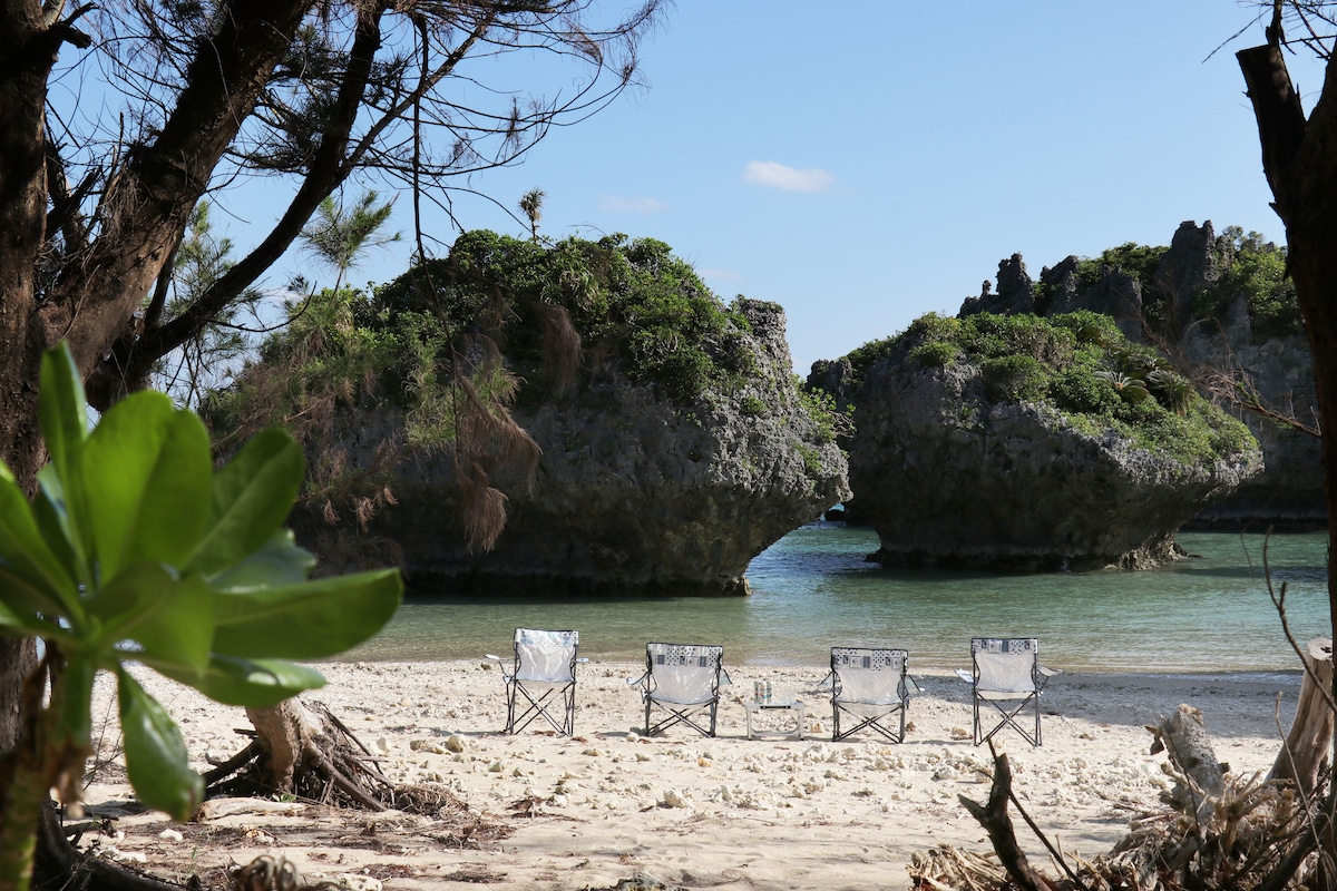 A serene beach scene features four white beach chairs arranged on soft sand, facing a calm turquoise lagoon. Two large, rugged rock formations rise from the water in the background, surrounded by lush greenery. The bright sky creates a peaceful atmosphere for relaxation.