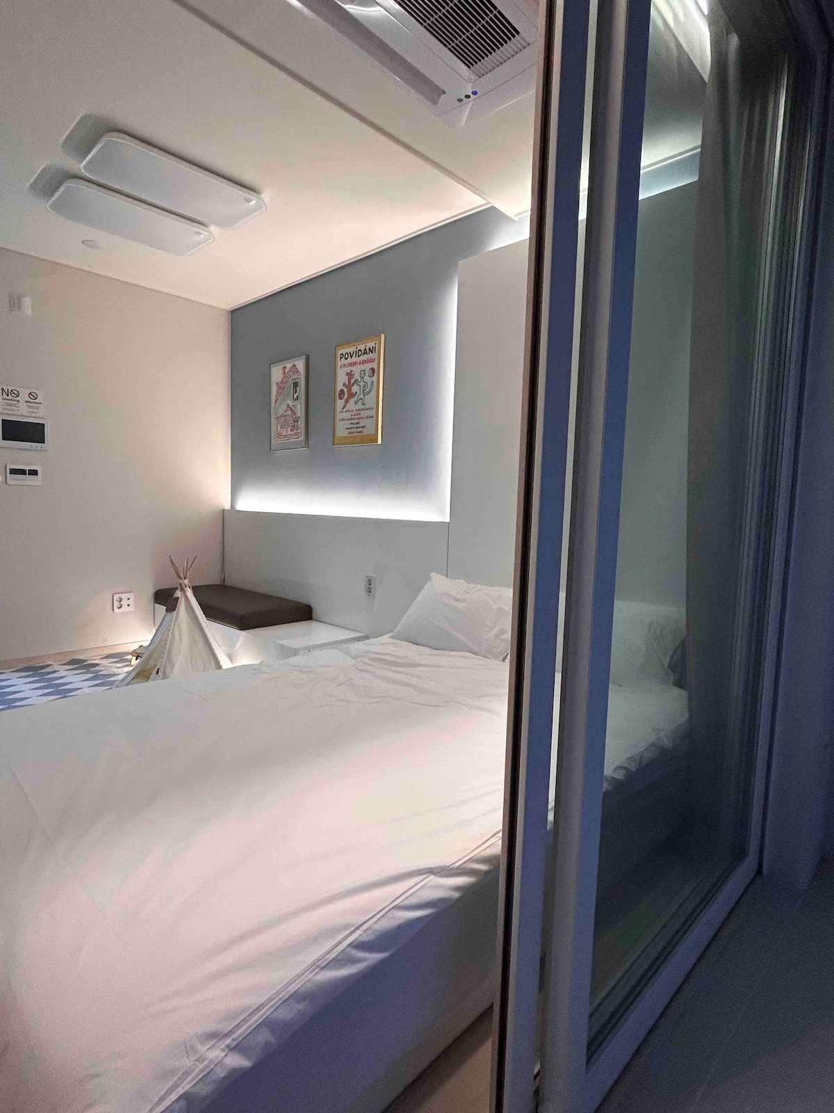A modern bedroom features a spacious bed covered with white linens. Wall art is displayed above a cozy seating area, and a lighted ambience enhances the overall feel. A glass partition separates the sleeping area from a small lounge space with a soft rug.