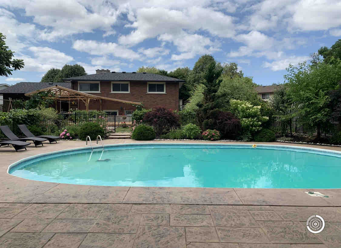 A private swimming pool is surrounded by well-maintained gardens and mature trees. Lounge chairs are positioned nearby for relaxation. The pool features steps leading into the water, while a covered seating area is partially visible, inviting outdoor enjoyment.