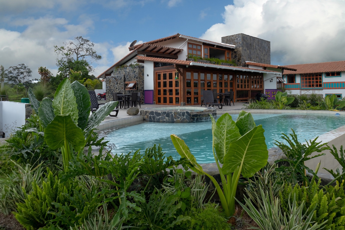 A spacious outdoor area features a private swimming pool, outlined by lush greenery and vibrant plants. The two-story chalet is visible in the background, showcasing wooden doors and stone accents beneath a partly cloudy sky.