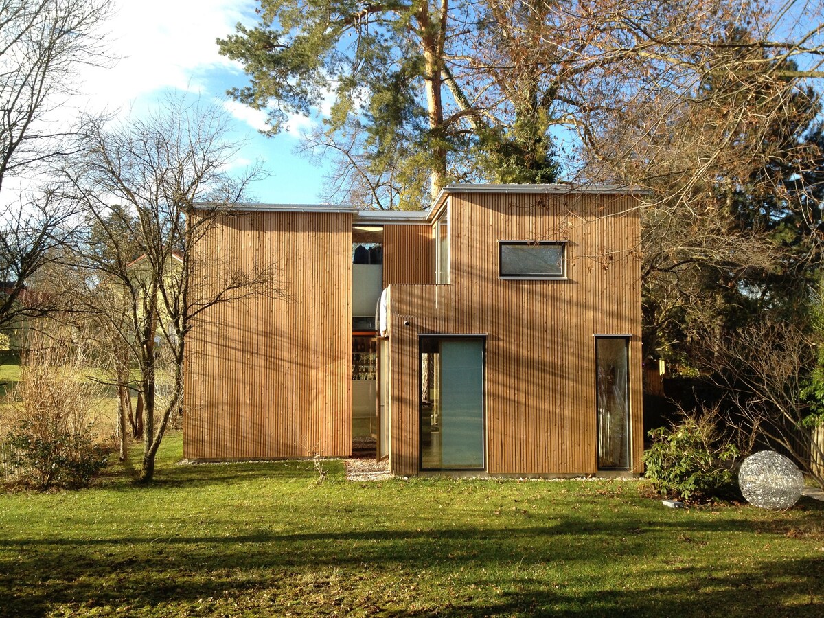 A modern wooden house is shown, featuring a distinctive façade with large windows and varied architectural elements. The structure is surrounded by greenery, including trees and grass, creating a serene outdoor environment. Natural light casts interesting shadows on the exterior.