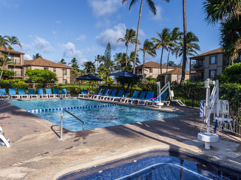 A sunny swimming pool is surrounded by lush tropical landscaping. Lounge chairs are arranged neatly around the pool, while umbrellas provide shade. Nearby, a hot tub is visible, offering a relaxing area. Tall palm trees sway gently in the background, enhancing the tropical ambiance.