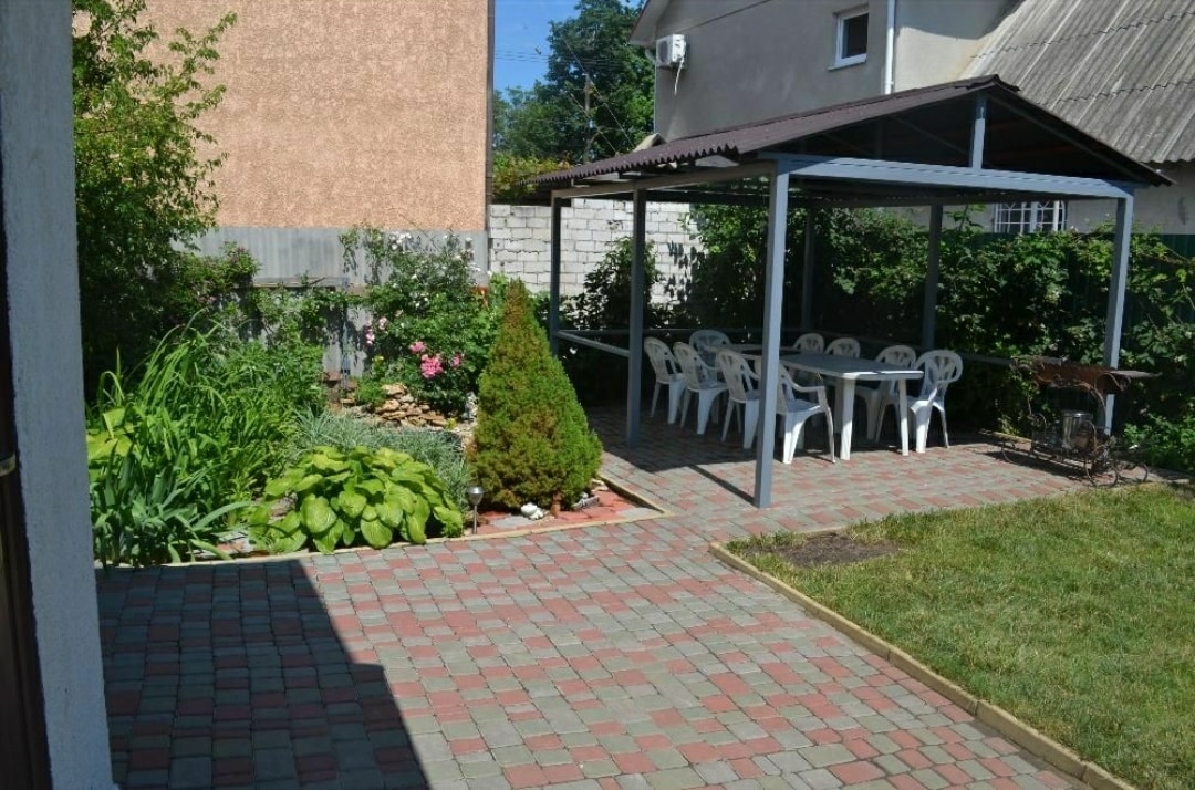 A shaded outdoor area features a large covered gazebo surrounded by a paved patio. Several white plastic chairs and a table are arranged underneath, with greenery and colorful flowers providing a natural backdrop. A decorative pond and manicured lawn are visible nearby.