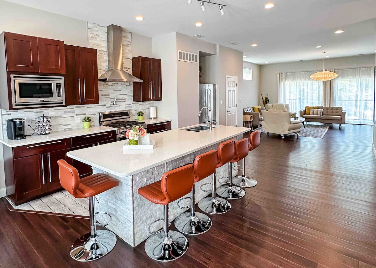 A modern kitchen showcases a large central island with six bar stools, featuring a sleek countertop and contemporary cabinetry. Stainless steel appliances and a decorative backsplash complement the space. The open layout connects seamlessly to a comfortable living area with large windows allowing natural light.