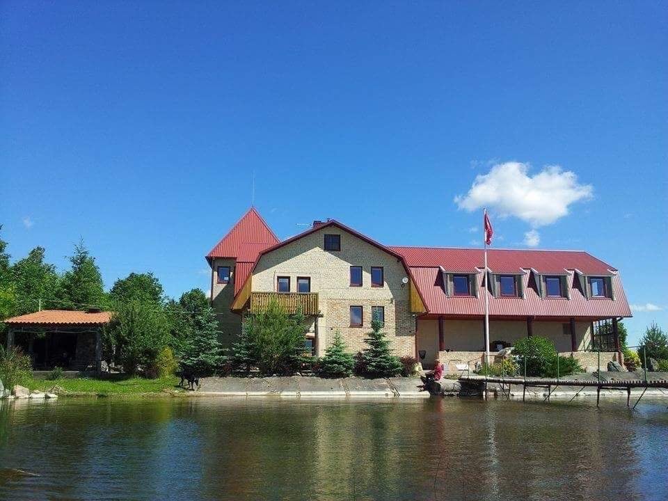 The exterior of a large building with a red-roofed design is shown, situated beside a calm body of water. Lush greenery surrounds the property, with various trees and shrubs visible. A flag is displayed in front, and there is a covered area to the left.