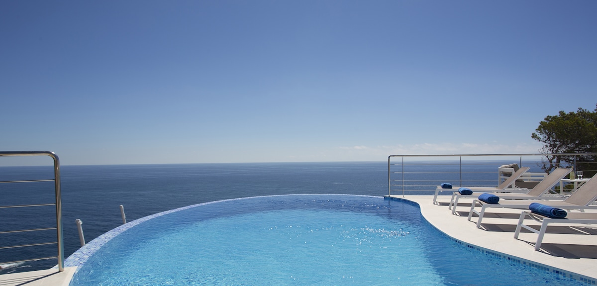 A kidney-shaped private pool is shown with clear blue water reflecting the sky. Sun loungers are arranged around the pool, overlooking a vast ocean view under a cloudless blue sky. The serene landscape enhances the tranquil atmosphere.