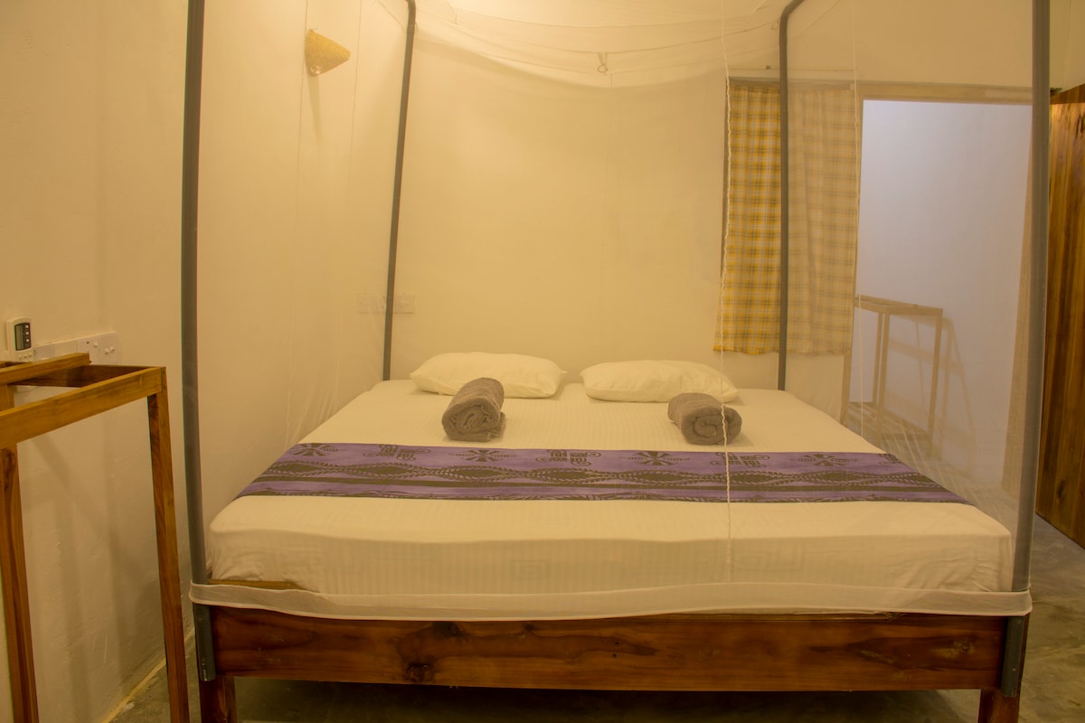 A spacious double bed is framed by a mosquito net, providing a serene sleeping environment. Soft gray towels are neatly arranged on the bed, which is accented by a vibrant patterned throw. Natural light filters in through a nearby window adorned with a checkered curtain.