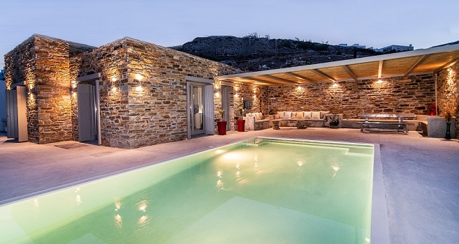 A spacious outdoor area is surrounded by stone walls, featuring a serene swimming pool with soft, illuminated water. Comfortable seating arrangements are present under a wooden roof, offering an inviting space for relaxation as the evening sky begins to darken.