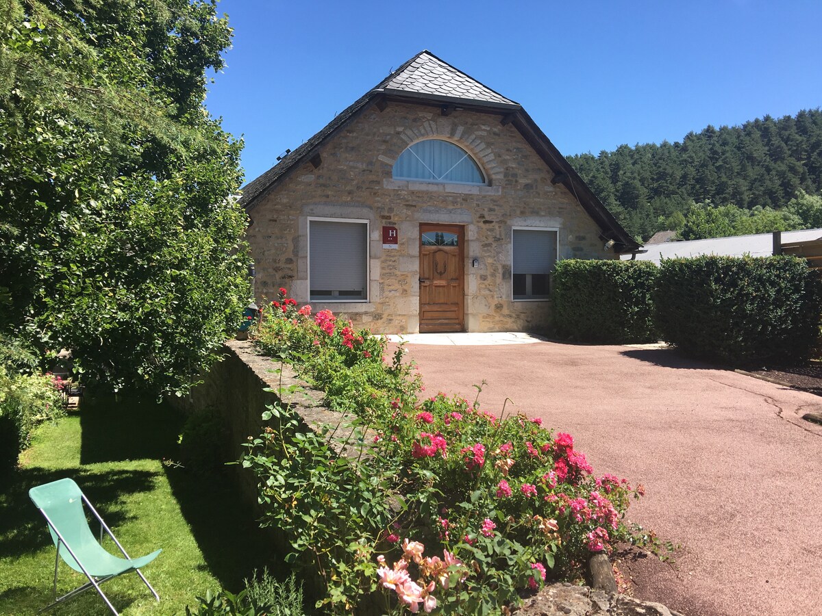 A charming stone house features a triangular roof and a welcoming front door. Surrounding gardens include green lawns and blossoming flowers, while two large windows provide ample natural light. The clean exterior is complemented by a well-maintained pathway and lush greenery, enhancing the inviting atmosphere.