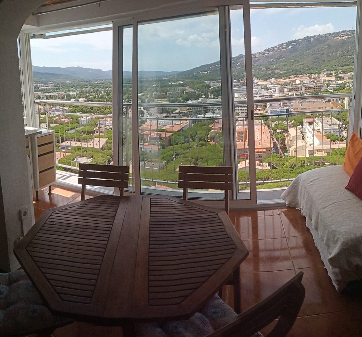 A spacious room features a round wooden table surrounded by wooden chairs. Large glass windows offer panoramic views of the mountainous landscape and the sea, enhancing the natural light that fills the area. A comfortable sofa bed is visible in the corner, adding to the inviting setting.