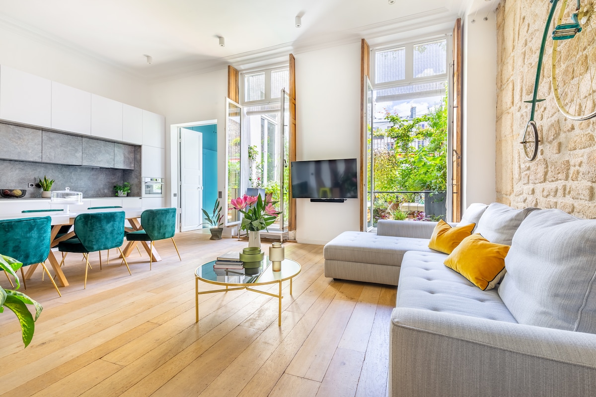 A spacious living area features a light grey couch with colorful cushions and a modern coffee table. Large windows allow natural light to fill the room, showcasing potted plants and greenery outside. A dining space is visible, with teal chairs arranged around a sleek table.