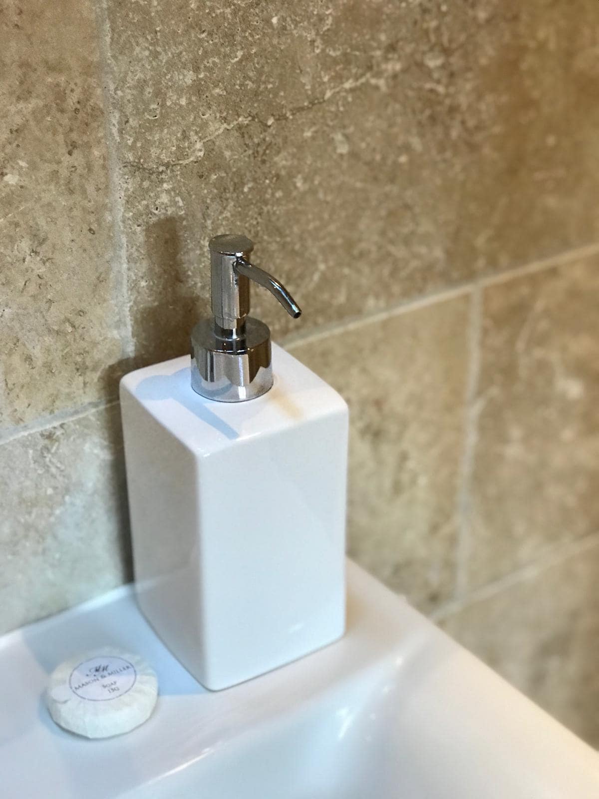 A sleek white soap dispenser is positioned on the edge of a light-colored sink. Beside it, a round, white soap bar is resting, with a subtle design on its surface. The background features textured stone tiles in warm tones, enhancing the bathroom's serene appearance.