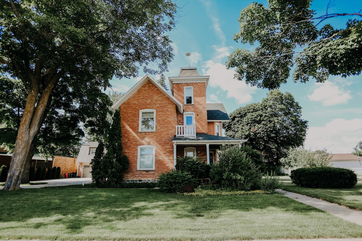 The exterior of a vintage brick home is showcased, featuring a tower and a small balcony. Lush green lawns surround the property, with mature trees providing shade. The sky is clear, and patches of clouds are visible, complementing the inviting atmosphere of the residence.