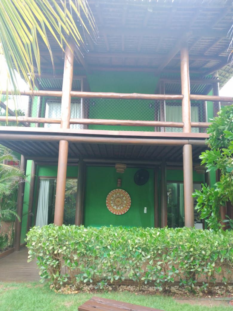 The exterior of a two-story structure features natural wood beams and vibrant green walls. A decorative wall piece adds character to the entrance, surrounded by lush greenery and a neatly trimmed hedge. Large windows allow for ample light, enhancing the inviting facade.