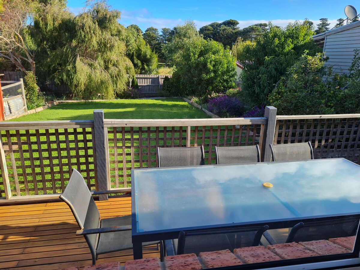 An outdoor dining area is positioned on a wooden deck, featuring a large glass table surrounded by several black chairs. Lush greenery is visible in the expansive backyard, creating a serene environment complemented by trees and blooming plants.