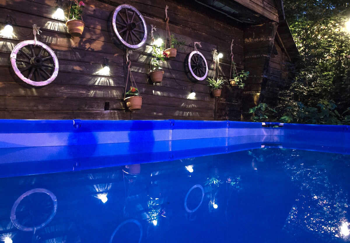 A blue pool reflects the lights from the surrounding area, creating a serene environment. Wooden wheels mounted on the wall add a rustic touch, while potted plants bring a hint of greenery to the scene.