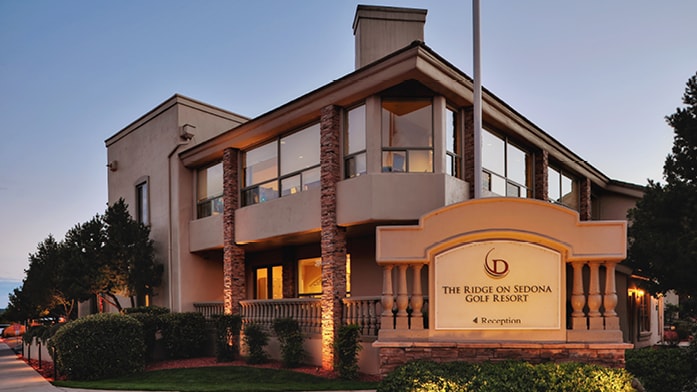 A welcoming resort entrance is highlighted by a large sign displaying the name 'The Ridge on Sedona Golf Resort.' The building features multiple large windows, with stone accents and lush greenery framing the facade. Soft exterior lighting enhances the evening ambiance.