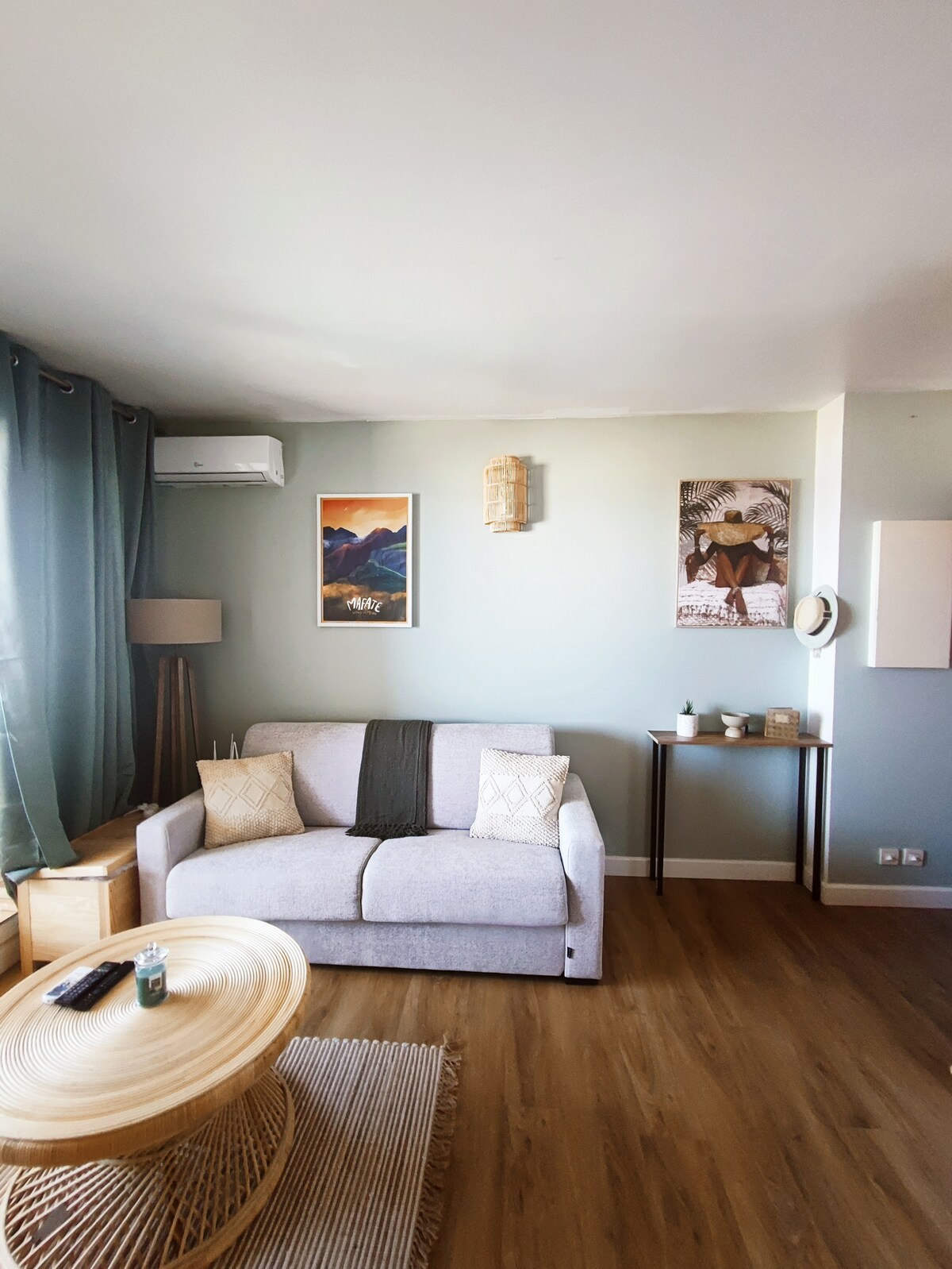 A comfortable living area features a light gray sofa adorned with decorative cushions. A coffee table made of natural materials sits in front. The walls are painted in soft green, complemented by artwork showcasing mountain landscapes. Ambient light filters in through large windows, accentuated by sheer curtains.