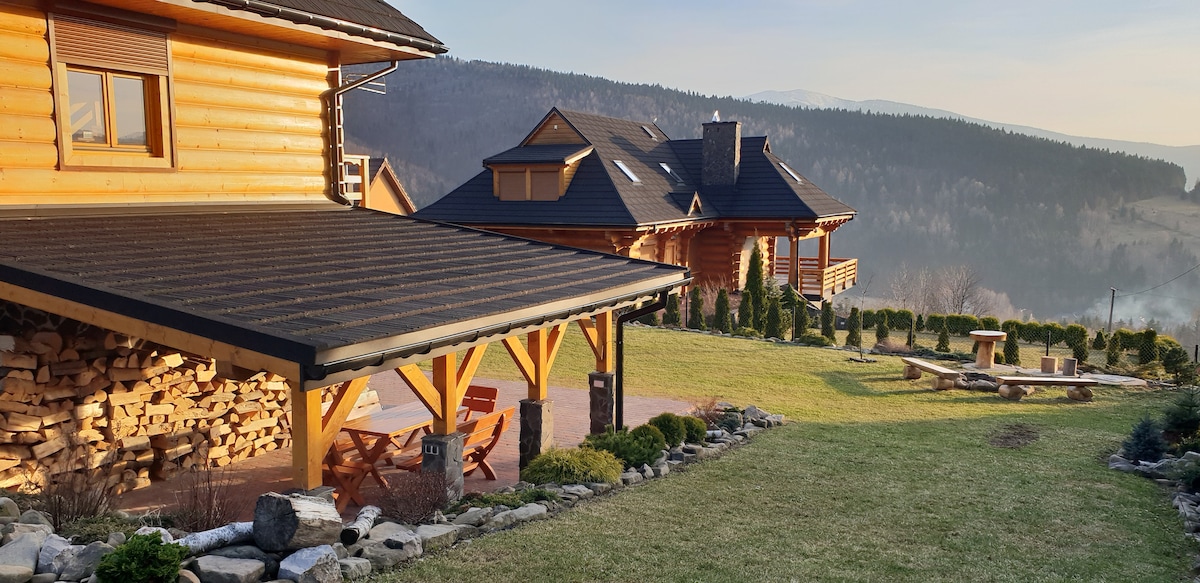 A wooden house is prominently featured with a covered porch, showcasing stacked firewood. The well-maintained garden is adorned with neatly trimmed hedges and a stone pathway, while a seating area is visible, inviting relaxation amid the scenic mountainous backdrop.