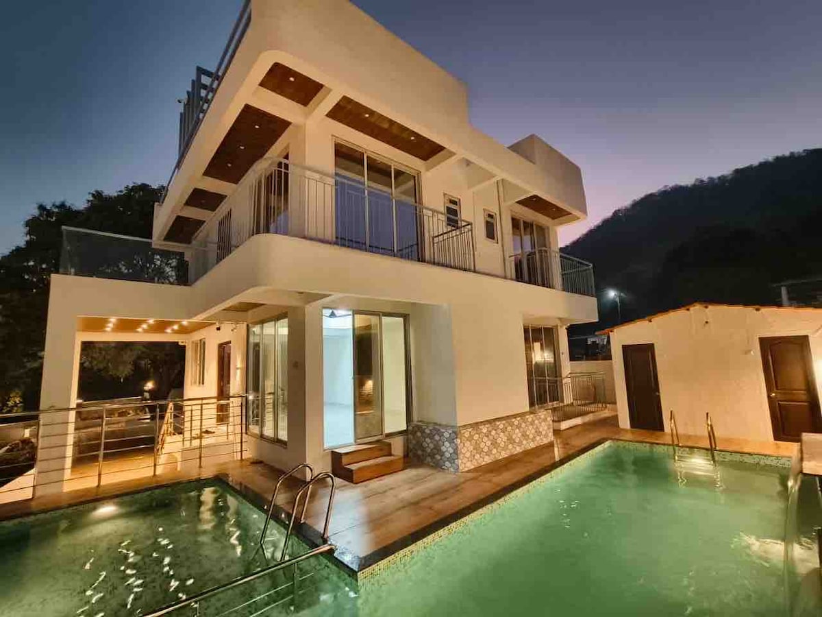 A modern two-story villa is visible, featuring large glass windows and a spacious balcony. Two private swimming pools are located at the front, surrounded by wooden decking. Soft evening light casts a warm glow over the structure, enhancing its contemporary design.