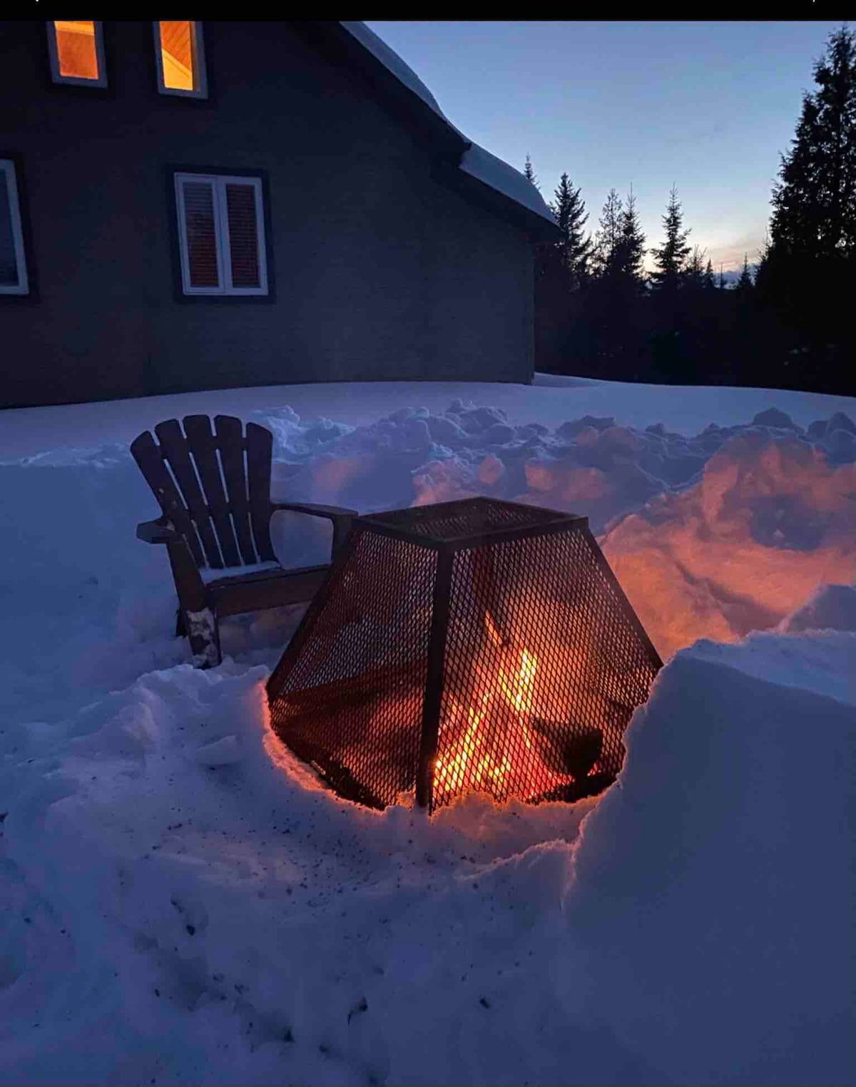 A fire pit is surrounded by snow, emitting a warm glow. A single wooden chair is positioned nearby. The backdrop features a house with multiple windows, partially illuminated by evening light, and evergreen trees are visible in the distance.
