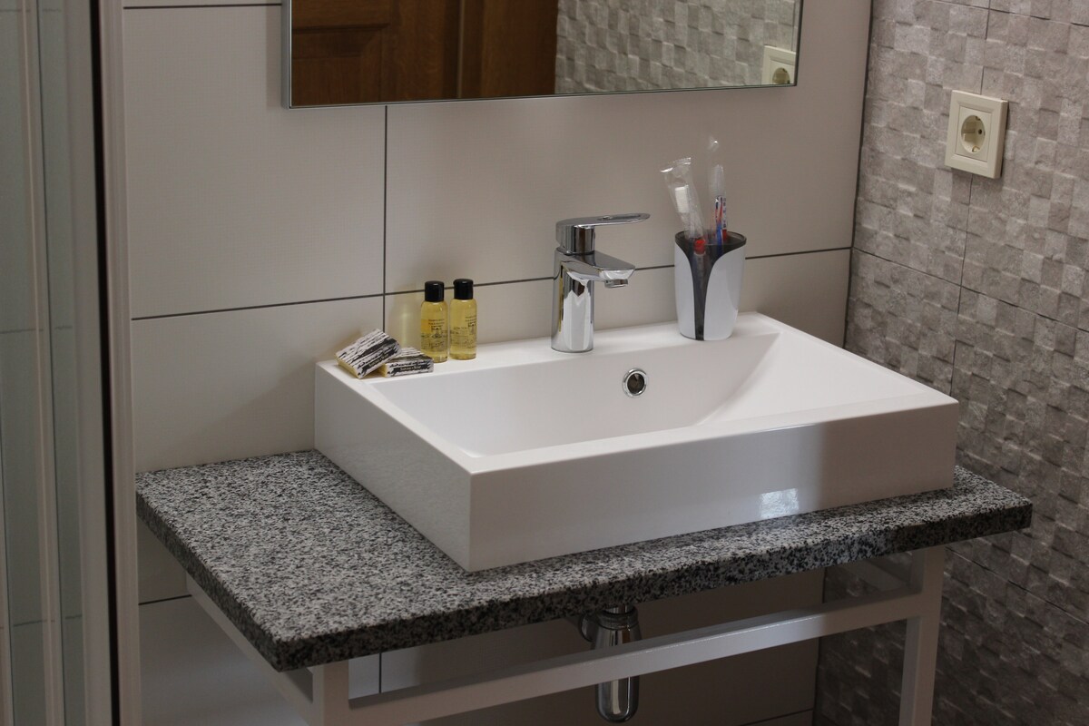 A modern bathroom sink is set on a granite countertop, complemented by a sleek faucet. Small toiletries are arranged beside the sink, and a mirror hangs above, reflecting the soft lighting and neutral tones of the tiled walls.