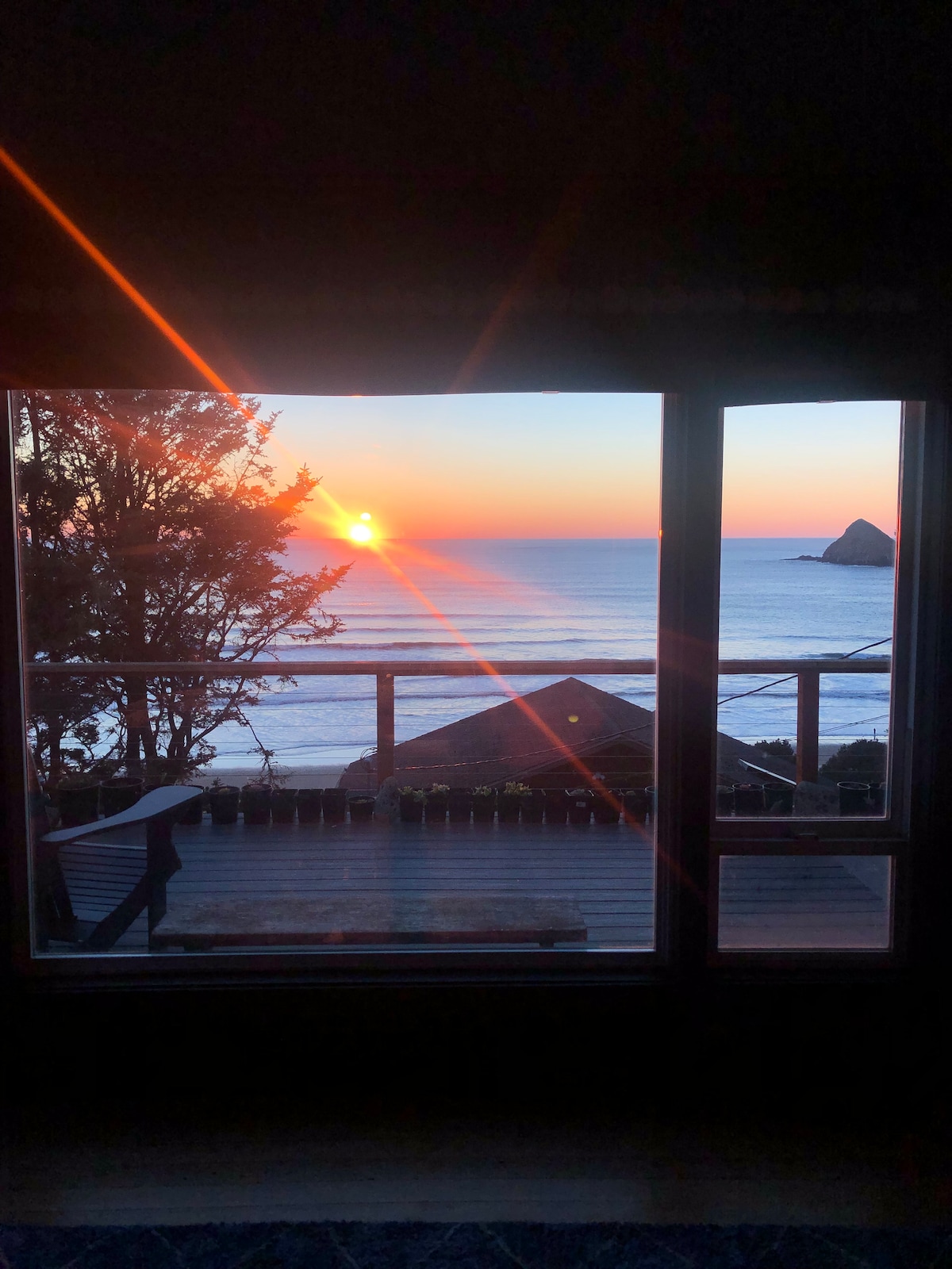 A view of a vibrant sunset over the Pacific Ocean is framed by large glass doors. The sun casts warm rays as it sinks into the horizon, illuminating the water and distant coastal rock formations. A wooden deck is visible in the foreground, surrounded by greenery.