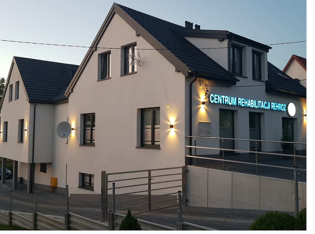 A modern rehabilitation center is presented, featuring a two-story building with a sleek white facade. Outdoor lighting emphasizes the entrance sign, 'Centrum Rehabilitacji Rehoz,' while adjacent parking and manicured landscaping add to the functional environment.