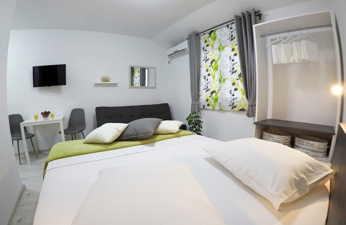 An inviting room features a spacious bed with multiple pillows. A sofa sits against the wall, while a flatscreen TV is mounted nearby. A small dining table with two chairs is positioned beside a window adorned with patterned curtains. Storage options are available in the closet.