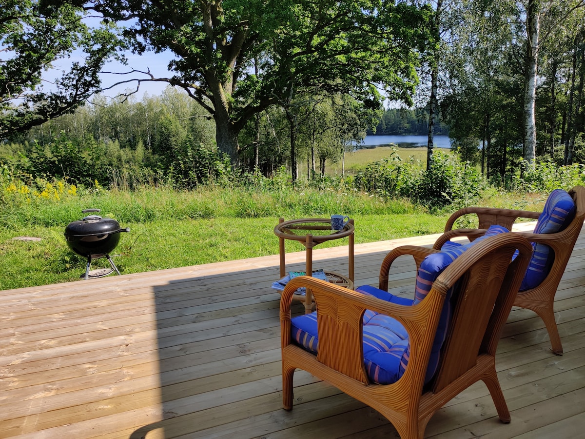 A wooden deck features two comfortable chairs with striped cushions and a small table, offering a scenic view of the lush green landscape and a nearby lake. A charcoal grill is positioned nearby, ready for outdoor cooking.