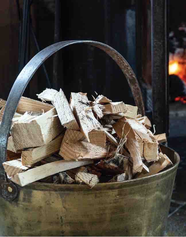 A brass bucket filled with neatly stacked firewood is positioned near a log burner. The warm glow of the flames can be seen in the background, creating a cozy ambiance.
