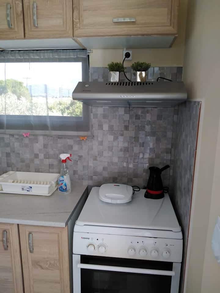 A compact kitchen features a stove and oven beneath a modern exhaust hood. A kettle is placed on the countertop next to a bottle of cleaning solution and a dish drying rack. Natural light is filtered through a window covered with sheer curtains.