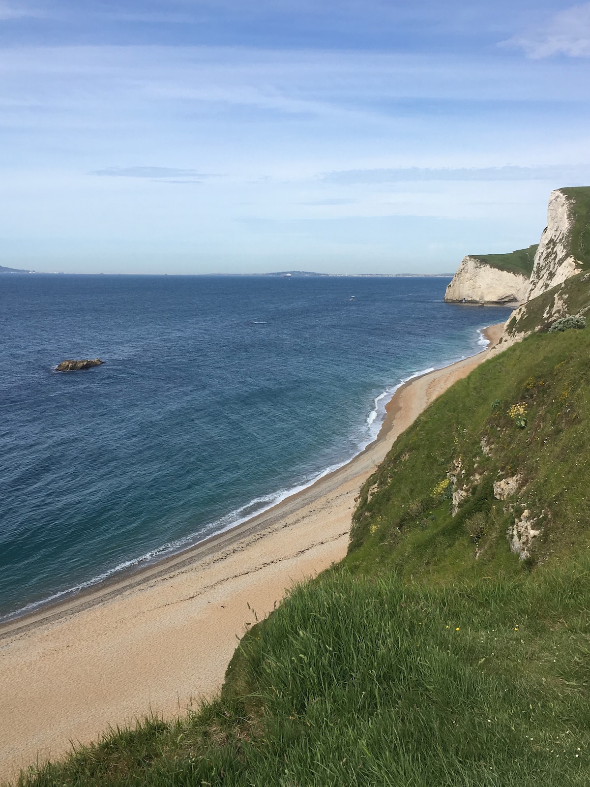 A serene coastal scene displays a sandy beach bordered by gentle waves, with lush green cliffs rising above. The clear water transitions into the distant horizon, featuring soft, rolling waves. A rocky formation is visible in the ocean, complementing the beautiful landscape.
