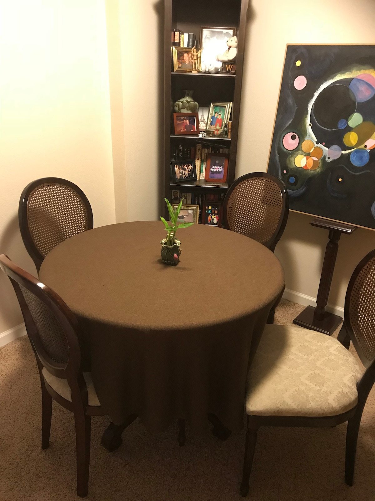 A round table with a brown tablecloth is set in a corner of the room, accompanied by four wooden chairs. A small plant sits at the center of the table. A bookshelf filled with various books and a colorful abstract painting adorn the wall behind.