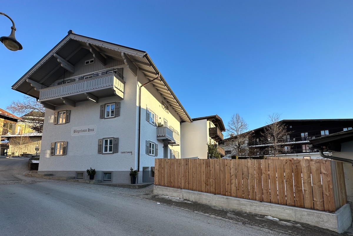 The exterior of a traditional-style building is showcased, featuring a peaked roof and multiple balconies. Light-colored walls contrast with darker window frames, creating a harmonious look. Surrounding structures and a wooden fence are visible, highlighting the building's location in a quaint alpine setting.