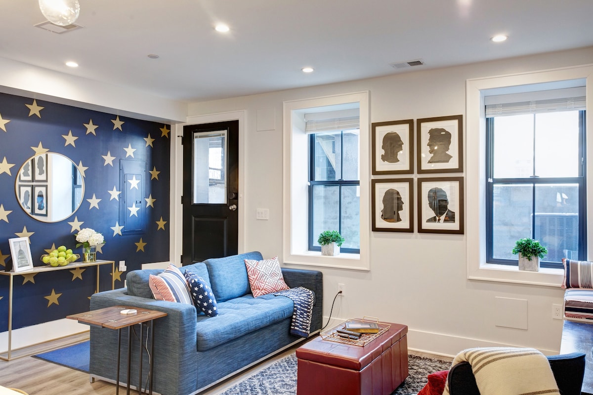 A living area features a blue sofa adorned with colorful pillows, complemented by a rustic coffee table. The wall displays decorative star patterns and framed silhouettes. Large windows provide natural light, and a door leads to the private entrance.