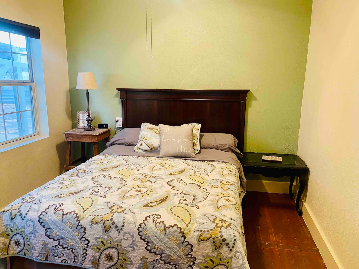 A cozy sleeping area is presented with a dark wood headboard and a patterned blanket covering the spacious bed. A bedside table holds a lamp, and natural light enters through the nearby window, enhancing the gentle green wall.