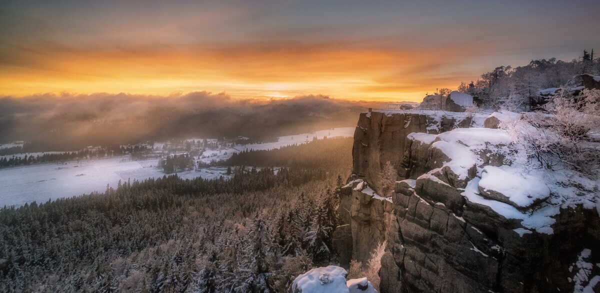 A winter landscape captures a dramatic cliffside overlooking a snow-covered valley. Soft morning light creates a warm glow across the horizon, while a blanket of clouds hovers above the serene wilderness, accentuating the natural beauty of the surroundings.