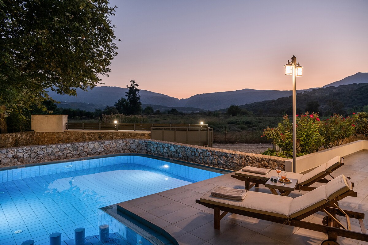 A spacious outdoor area features a heated pool with a soothing blue glow, surrounded by modern sunbeds. Lush greenery and a serene mountain view provide a tranquil backdrop, while gentle lighting adds to the peaceful atmosphere at dusk.