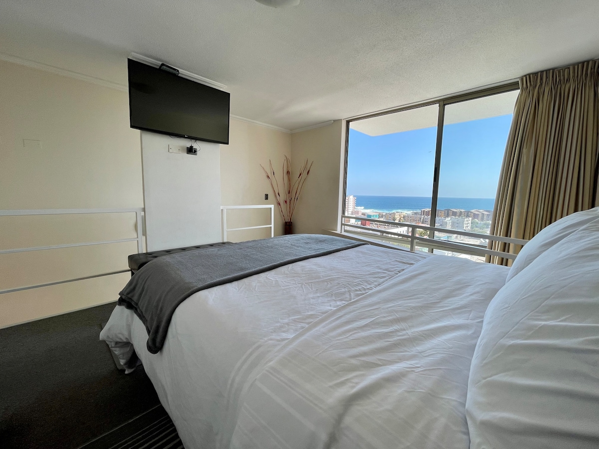 A spacious bedroom features a large bed with crisp white linens and a gray throw blanket. A wall-mounted TV is positioned in front of the bed, while large windows provide an expansive view of the ocean and city skyline.