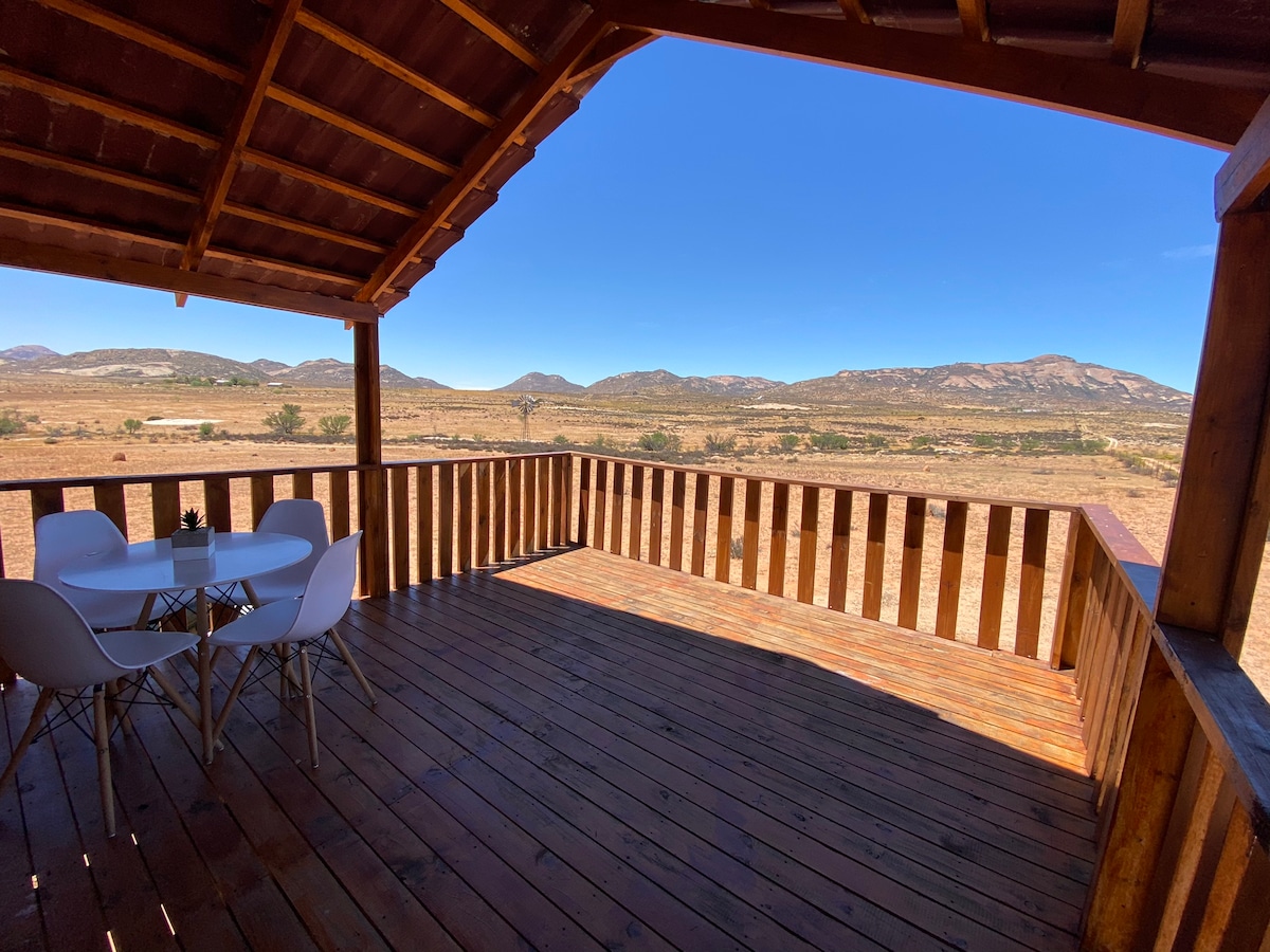 A spacious wooden deck is presented with a dining table and chairs, allowing for outdoor seating. The surrounding landscape features rolling hills and a clear blue sky, creating an open and serene environment.
