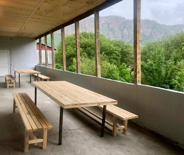 A spacious terrace features wooden tables and benches, allowing for communal dining. Large windows provide scenic views of the surrounding mountains and greenery, enhancing the outdoor ambiance. The area is sheltered under a wooden roof, offering comfort in various weather conditions.