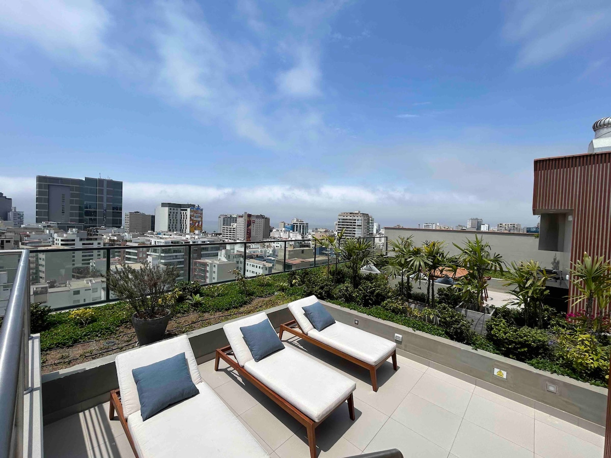 An outdoor terrace features stylish lounge chairs arranged on a tiled surface, surrounded by greenery and potted plants. A panoramic city view is visible in the background, showcasing a mix of modern buildings and open skies.