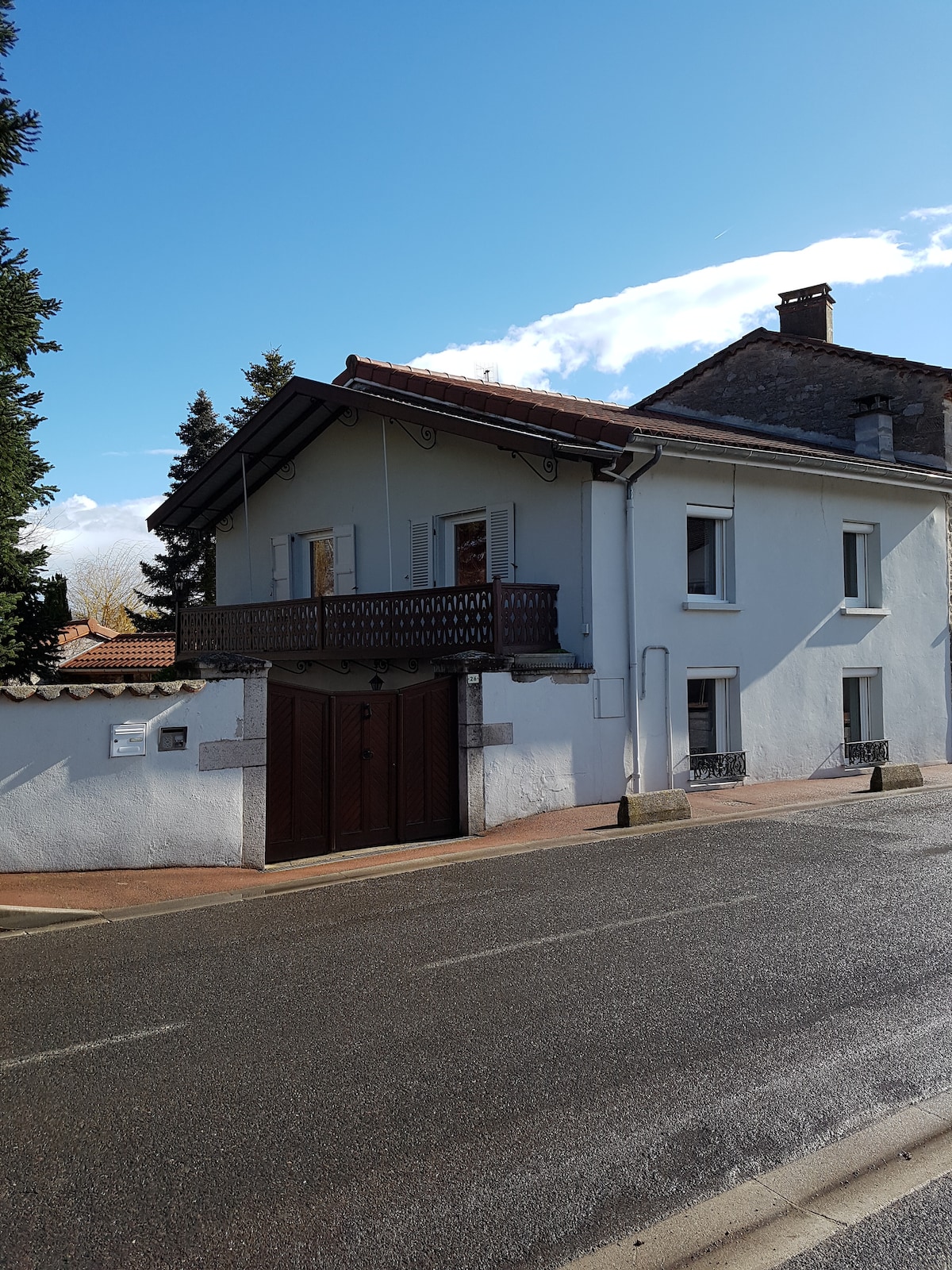 A two-story white house is framed by trees and a clear blue sky. The building features a wooden balcony on the upper level, while large gates provide access to the entrance. The exterior is simple, with traditional architectural elements visible.