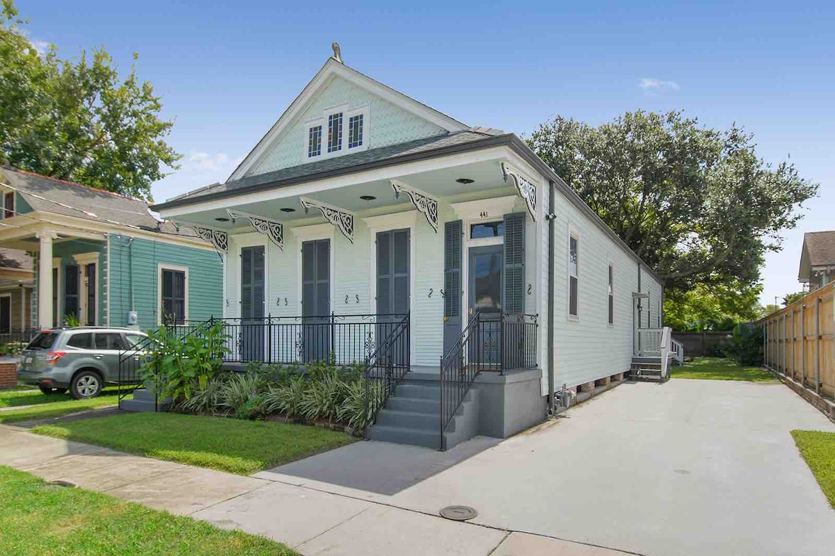 A charming historic home is featured, showcasing a light blue exterior with white trim and decorative window awnings. A spacious driveway provides parking space, with well-maintained landscaping lining the entrance. The building is situated in a peaceful neighborhood, surrounded by trees and other residential homes.